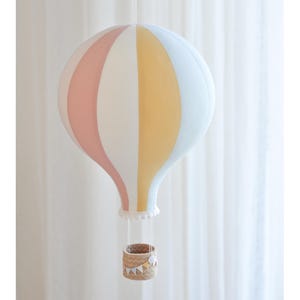 Può includere: Un pallone aerostatico bianco, rosa e giallo con un cesto di vimini appeso. Il pallone è decorato con un bordo di pizzo bianco e piccole bandierine.