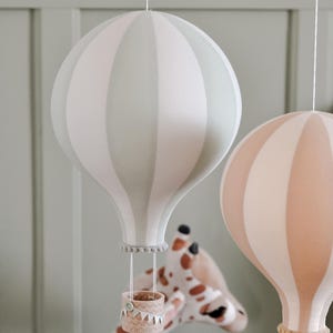 May include: Two decorative hot air balloon mobiles in soft pastel colors. One is striped in light green and white, the other in peach and white. Each has a small basket and hangs from a string. A giraffe plush toy is visible in the background.