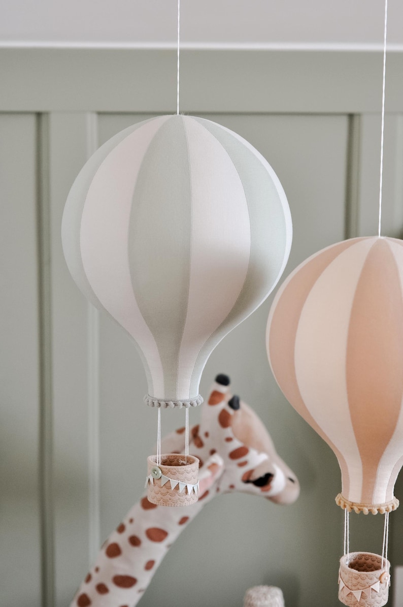 May include: Two decorative hot air balloon mobiles in soft pastel colors. One is striped in light green and white, the other in peach and white. Each has a small basket and hangs from a string. A giraffe plush toy is visible in the background.