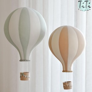 May include: Two decorative hot air balloon hanging lamps. One is light green and white striped, the other is peach and white striped. Each has a woven basket and is suspended by strings. The lamps are against a white curtain background.