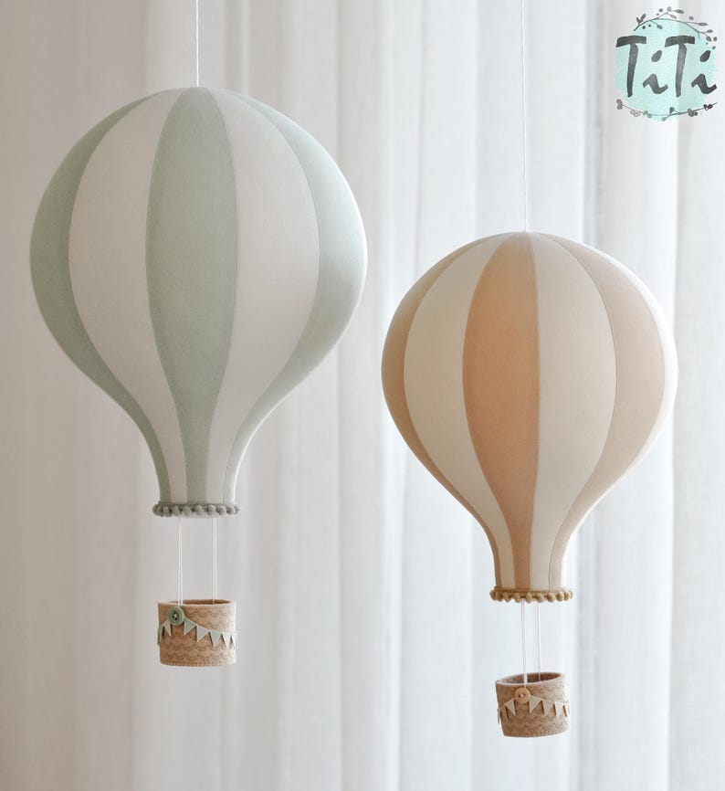 May include: Two decorative hot air balloon hanging lamps. One is light green and white striped, the other is peach and white striped. Each has a woven basket and is suspended by strings. The lamps are against a white curtain background.
