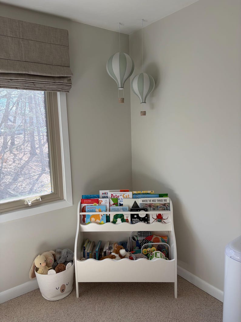 May include: A children's room corner featuring a white bookshelf filled with books and toys. Two decorative hot air balloon lights hang from the ceiling. A white basket with a bear face design holds stuffed animals. The room has a neutral color palette.