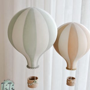 May include: Two decorative hot air balloon mobiles. One is light green and white, the other is tan and white. Each has a woven basket and is suspended by a string. The balloons are striped and have a whimsical, nursery-like aesthetic.