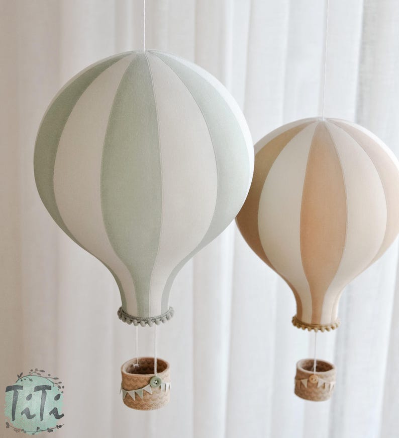 May include: Two decorative hot air balloon mobiles. One is light green and white, the other is tan and white. Each has a woven basket and is suspended by a string. The balloons are striped and have a whimsical, nursery-like aesthetic.