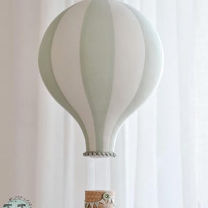 May include: A decorative hot air balloon mobile in soft green and white stripes. The balloon hangs from a string, with a small basket below. The basket is decorated with a scalloped edge and a button.