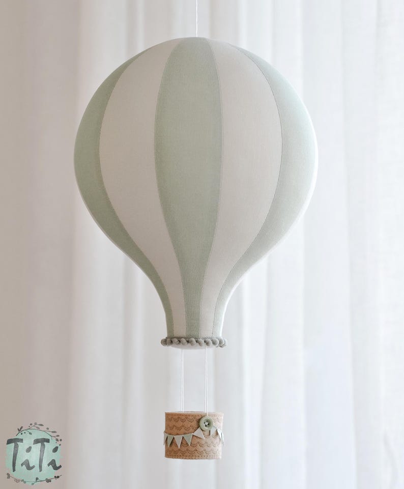 May include: A decorative hot air balloon mobile in soft green and white stripes. The balloon hangs from a string, with a small basket below. The basket is decorated with a scalloped edge and a button.