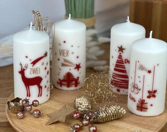 May include: Four white pillar candles with red and gold Christmas designs, including a reindeer, Christmas tree, and gifts. The candles are on a wooden tray with decorative ornaments and a star.