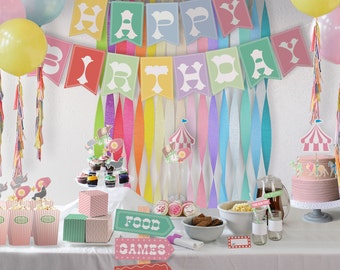 May include: A colorful birthday party table setting with a circus theme. The table is decorated with a banner that says "Happy Birthday", colorful streamers, and a pink cake with a circus tent topper. There are also cupcakes, popcorn, and other treats on the table. The table is set with signs that say "Food", "Games", "Attractions", and "Restrooms".