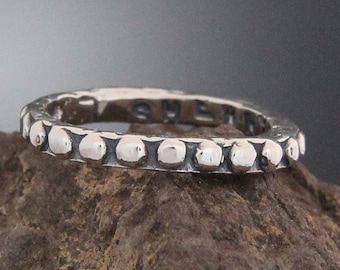 Könnte beinhalten: Ein silberner Ring mit einer Reihe kleiner, erhabener, kreisförmiger Designs um das Band. Der Ring liegt auf einem rauen, braunen Stein. Das Wort "SHE" ist auf der Innenseite des Rings sichtbar.