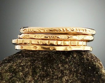 May include: A stack of five gold-colored rings with etched designs. The rings are stacked on a dark, textured surface, with a gradient gray background. The rings appear to be made of metal.