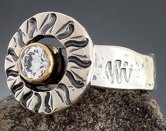 Puede incluir: Un anillo de plata con un diseño de sol alrededor de una piedra preciosa redonda y transparente engastada en un bisel dorado. La banda del anillo tiene un patrón texturizado. El anillo se muestra sobre una superficie rocosa texturizada.
