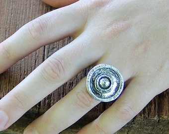 May include: A silver-toned ring with a circular design featuring concentric circles and a central raised element. The ring is worn on a hand resting on a wooden surface. The ring's design is detailed and textured.