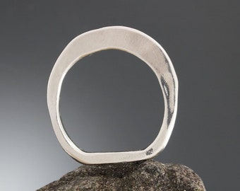 Könnte beinhalten: Ein silberner Ring mit organischer, asymmetrischer Form. Der Ring besteht aus einem dicken Metallband und hat eine glatte, polierte Oberfläche.