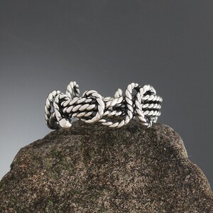 K&ouml;nnte beinhalten: Ein silberfarbener Ring mit Seil-Design. Der Ring besteht aus ineinander verschlungenen, seilartigen Str&auml;ngen, die eine strukturierte, nautische Optik erzeugen. Er wird auf einem rauen, grauen Stein vor einem dunkelgrauen Hintergrund pr&auml;sentiert.