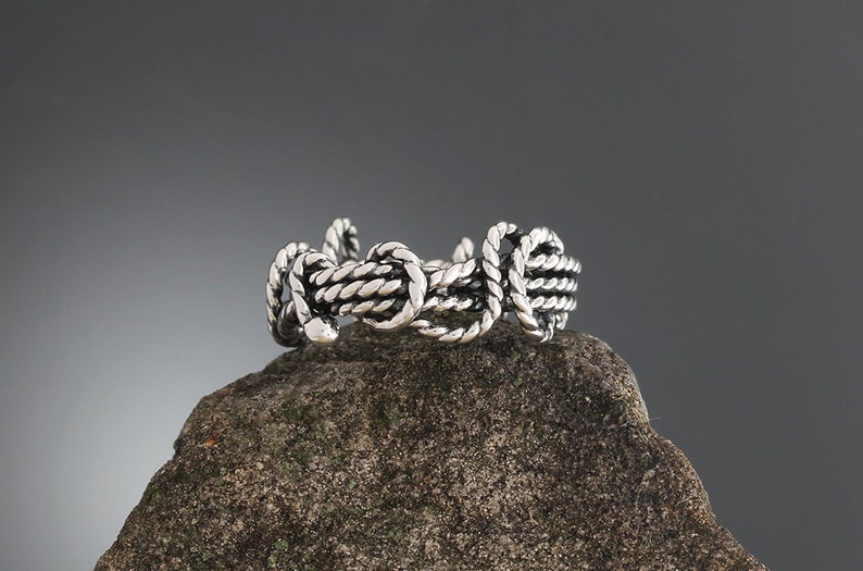 K&ouml;nnte beinhalten: Ein silberfarbener Ring mit Seil-Design. Der Ring besteht aus ineinander verschlungenen, seilartigen Str&auml;ngen, die eine strukturierte, nautische Optik erzeugen. Er wird auf einem rauen, grauen Stein vor einem dunkelgrauen Hintergrund pr&auml;sentiert.