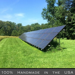 K&ouml;nnte beinhalten: Solarpanelanlage auf einer Wiese unter einem strahlend blauen Himmel. Die Paneele sind abgewinkelt, um Sonnenlicht einzufangen, mit B&auml;umen im Hintergrund. Der Text unten lautet "100% Handmade in the USA using solar energy."