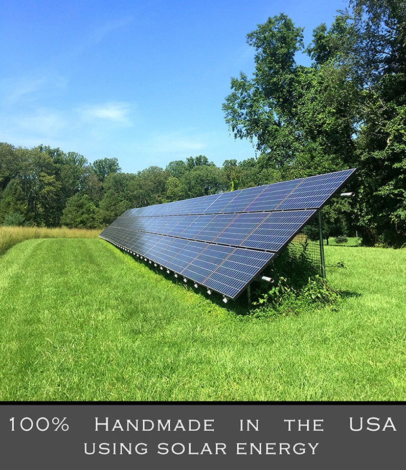 K&ouml;nnte beinhalten: Solarpanelanlage auf einer Wiese unter einem strahlend blauen Himmel. Die Paneele sind abgewinkelt, um Sonnenlicht einzufangen, mit B&auml;umen im Hintergrund. Der Text unten lautet "100% Handmade in the USA using solar energy."
