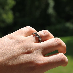 K&ouml;nnte beinhalten: Ein handgefertigter Ring mit einem einzigartigen, gewebten Design. Der Ring besteht aus dunklem Metall, m&ouml;glicherweise Bronze oder Kupfer, und weist ein kompliziertes Muster auf. Der Ring wird an einem Finger getragen und zeigt seine detaillierte Handwerkskunst. Der Hintergrund ist verschwommen, mit gr&uuml;nem Laub.