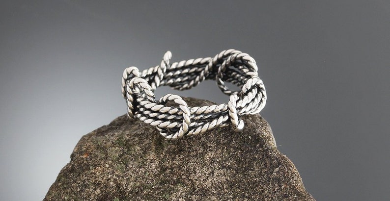 K&ouml;nnte beinhalten: Ein silberner Ring mit Seildesign. Der Ring besteht aus verdrehten Metallstr&auml;ngen, die ein strukturiertes Aussehen erzeugen. Er wird auf einem rauen, grauen Stein vor neutralem Hintergrund pr&auml;sentiert.