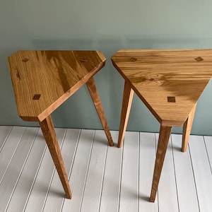 May include: Two triangular wooden side tables with a light brown wood finish and three legs each. The tables have a unique design with square inlays on the top.