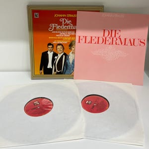 May include: A framed album cover and a pink album cover with the text "Die Fledermaus" and the name Johann Strauss. Two vinyl records are visible in white sleeves with red labels.