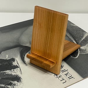 Puede incluir: Soporte de teléfono de madera de color marrón claro. El soporte tiene una parte trasera rectangular y una pequeña repisa en la parte inferior para sostener un teléfono. El soporte está inclinado para facilitar la visualización.