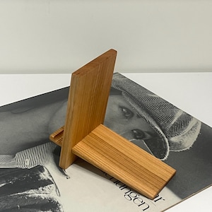 May include: A wooden phone stand with a rectangular base and a vertical support. The stand is made of light-colored wood and has a natural grain pattern. The stand is designed to hold a phone or tablet.
