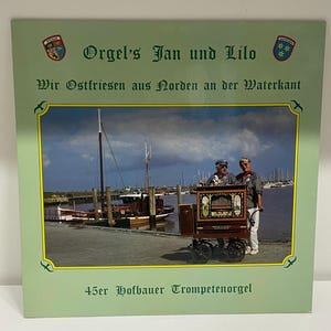May include: A vintage album cover with the text "Orgel's Jan und Lilo" and "Wir Ostfriesen aus Norden an der Waterkant." The image features a photo of two people with a street organ near a harbor. The text "45er Hofbauer Trompetenorgel" is also present.