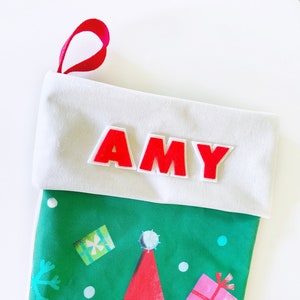 May include: A Christmas stocking with a white cuff and a red ribbon loop. The stocking is green with a snowman and gift pattern. The name "AMY" is in red letters on the cuff.
