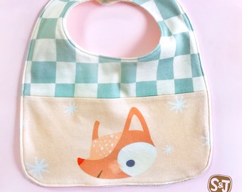 May include: A light blue and white checkered baby bib with a fox design and a pocket. The bib has a white button closure. The text "S&J Novelty" is printed on the bottom of the bib.