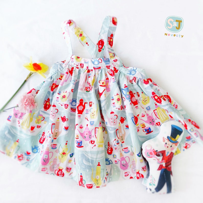 May include: A light blue sleeveless dress with a teacup and teapot pattern. The dress has adjustable straps and a gathered skirt. The dress is paired with a small toy doll wearing a top hat and a red vest.