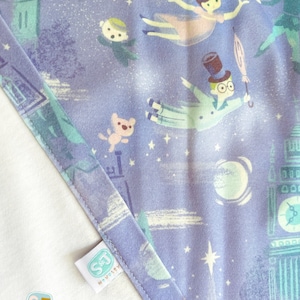 K&ouml;nnte beinhalten: Lilafarbener Stoff mit einem sich wiederholenden Muster aus einem fliegenden Jungen, einem M&auml;dchen, einem Teddyb&auml;r und einem Uhrturm. Der Stoff hat einen wei&szlig;en Rand und ein wei&szlig;es Etikett mit dem Text "S&J Novelty".