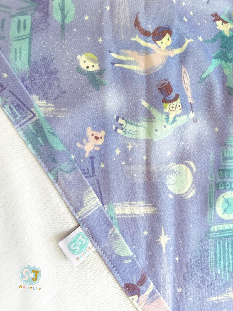 K&ouml;nnte beinhalten: Lilafarbener Stoff mit einem sich wiederholenden Muster aus einem fliegenden Jungen, einem M&auml;dchen, einem Teddyb&auml;r und einem Uhrturm. Der Stoff hat einen wei&szlig;en Rand und ein wei&szlig;es Etikett mit dem Text "S&J Novelty".