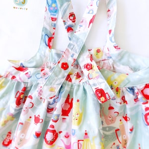 May include: A light blue sleeveless dress with a white and red teacup and teapot pattern. The dress has two straps that cross in the back and button at the back.