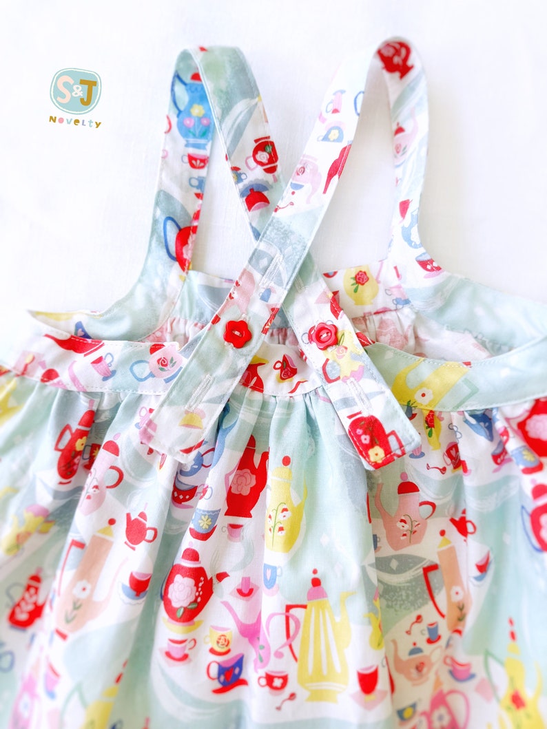 May include: A light blue sleeveless dress with a white and red teacup and teapot pattern. The dress has two straps that cross in the back and button at the back.