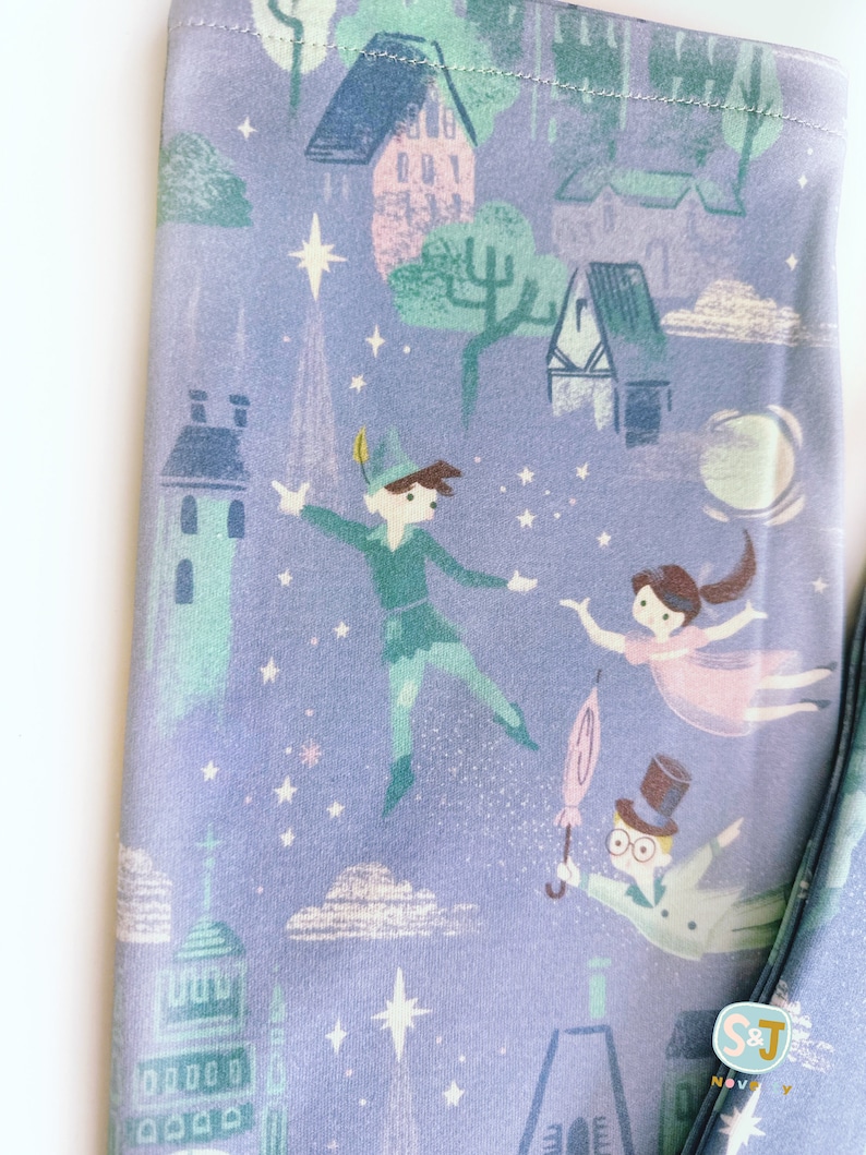 K&ouml;nnte beinhalten: Lilafarbener Stoff mit einem sich wiederholenden Muster eines Nachthimmels mit Sternen, Wolken, Geb&auml;uden und Figuren aus Peter Pan. Die Figuren sind Peter Pan, Wendy und Captain Hook.
