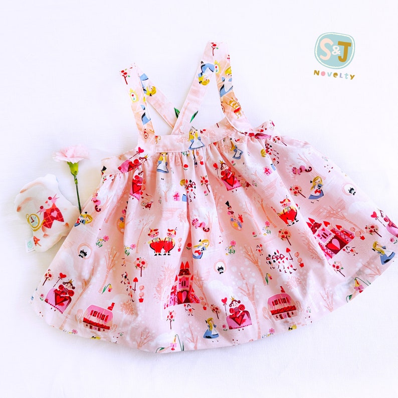 May include: A pink sleeveless dress with a criss-cross strap back. The dress has a whimsical print featuring characters and scenes from Alice in Wonderland. The dress is made of a soft, lightweight fabric.
