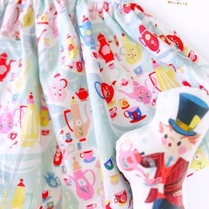 May include: A light blue dress with a white background and a pattern of teacups, teapots, and flowers. The dress is made of a soft, lightweight fabric and has a gathered skirt. The dress is paired with a plush doll of a character wearing a top hat and a red jacket.