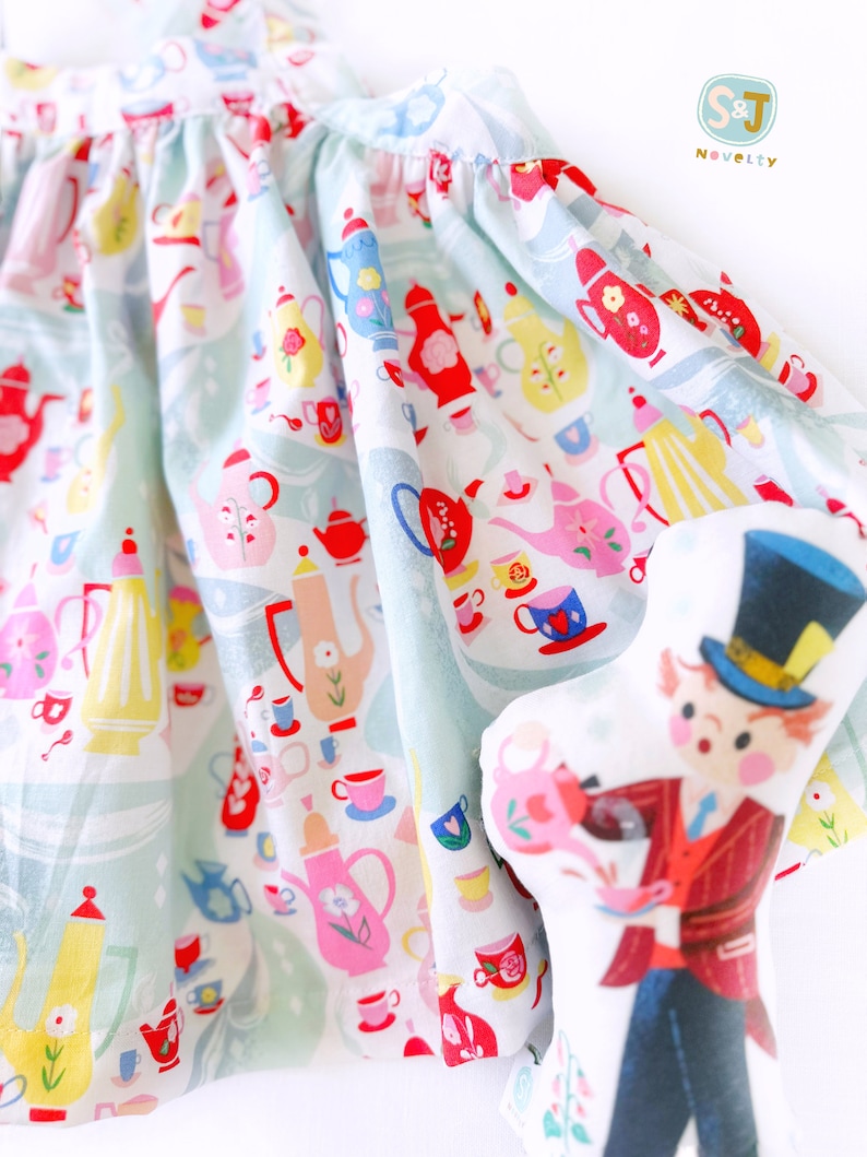 May include: A light blue dress with a white background and a pattern of teacups, teapots, and flowers. The dress is made of a soft, lightweight fabric and has a gathered skirt. The dress is paired with a plush doll of a character wearing a top hat and a red jacket.