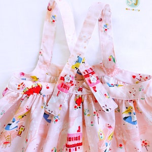 May include: Pink sleeveless dress with a white and pink floral print featuring Alice in Wonderland characters. The dress has adjustable straps with white buttons.
