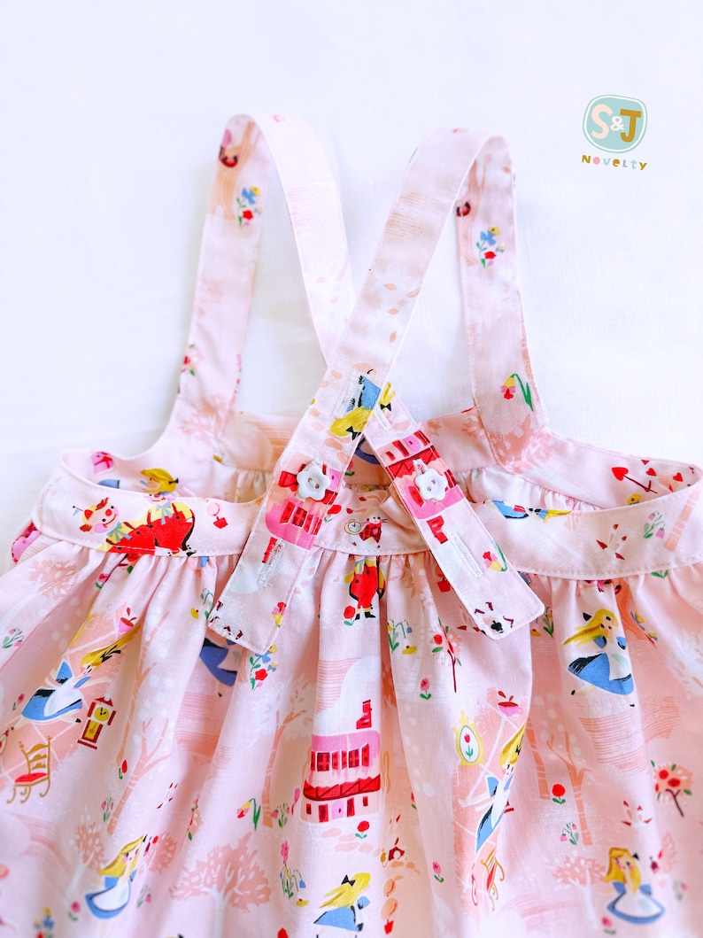 May include: Pink sleeveless dress with a white and pink floral print featuring Alice in Wonderland characters. The dress has adjustable straps with white buttons.