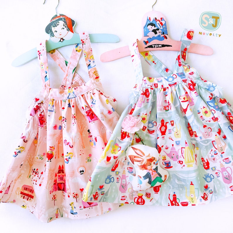 May include: Two pink and blue sleeveless dresses with a criss-cross strap back. The dresses feature a whimsical print of teacups, teapots, and other tea party elements. The dresses are hanging on wooden hangers with a vintage design.