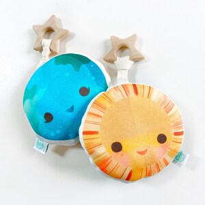 May include: Two plush baby toys: one blue Earth and one yellow Sun, each with a smiling face. They have wooden star-shaped teethers attached with white ribbon. The toys are on a white background.