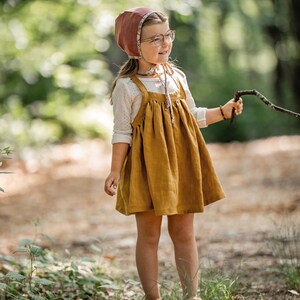 May include: A young girl wearing a brown and floral bonnet, a white long-sleeved shirt, and a mustard yellow linen dress with straps. She is standing in a forest setting and holding a twig.
