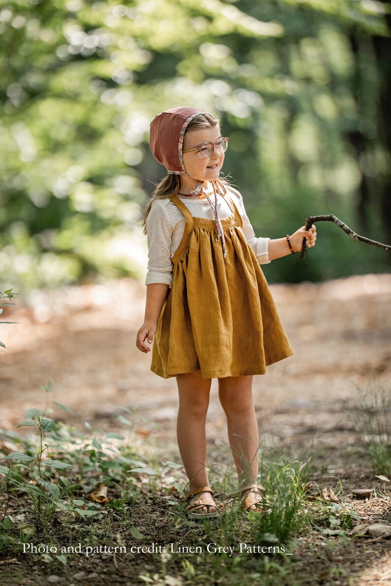 May include: A young girl wearing a brown and floral bonnet, a white long-sleeved shirt, and a mustard yellow linen dress with straps. She is standing in a forest setting and holding a twig.