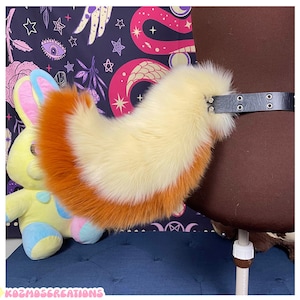 Puede incluir: Una cola de piel sintética esponjosa en crema y naranja, sujeta a un cinturón negro con herrajes plateados. La cola está diseñada para parecerse a una cola de zorro. Un peluche de conejito está en el fondo.