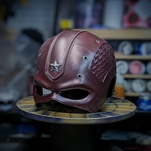 May include: A burgundy leather helmet with a star on the front and a textured design on the side. The helmet has a visor and is sitting on a turntable.