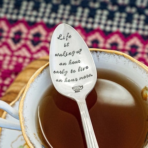 May include: A silver teaspoon with the inscription "Life is waking up an hour early to live an hour more." The spoon is resting in a white teacup with a gold rim and floral pattern. The teacup is on a wooden surface.