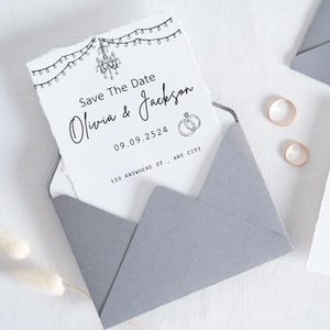 Puede incluir: Un sobre gris contiene una tarjeta blanca con el texto "Save The Date" y los nombres "Olivia & Jackson". La tarjeta presenta una ilustraci&oacute;n de una l&aacute;mpara de ara&ntilde;a y luces de cadena. Se ven dos anillos dorados.