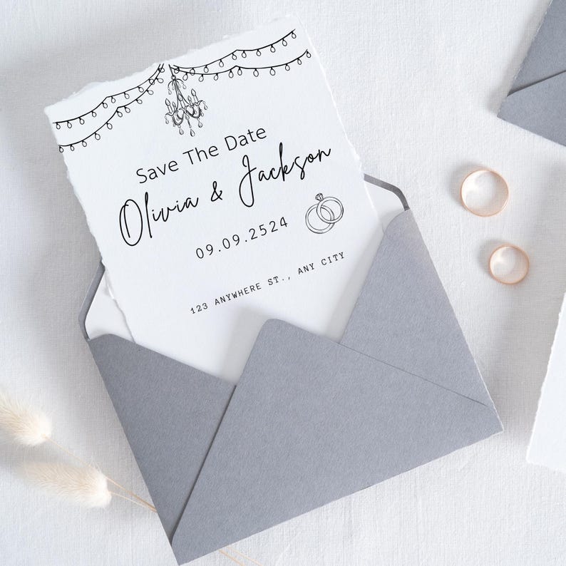 Puede incluir: Un sobre gris contiene una tarjeta blanca con el texto "Save The Date" y los nombres "Olivia & Jackson". La tarjeta presenta una ilustraci&oacute;n de una l&aacute;mpara de ara&ntilde;a y luces de cadena. Se ven dos anillos dorados.